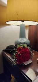 A bouquet of red roses with greenery is placed on a wooden nightstand beside a light blue vase-like lamp with a beige shade. The scene communicates a sense of elegance and coziness.