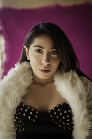 A person with long dark hair is wearing a black outfit embellished with gold studs and a white fur-trimmed garment. The background features a rich purple color, contributing to an elegant and sophisticated atmosphere.