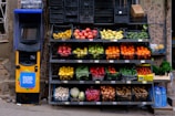A colorful display of various fruits and vegetables arranged in black crates outside a store, next to a yellow vending machine with signage in a foreign script. The produce includes apples, oranges, lemons, peppers, potatoes, and onions with some water bottles nearby.