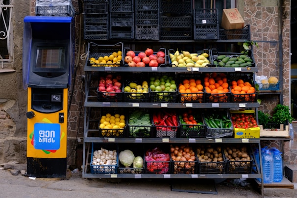 A colorful display of various fruits and vegetables arranged in black crates outside a store, next to a yellow vending machine with signage in a foreign script. The produce includes apples, oranges, lemons, peppers, potatoes, and onions with some water bottles nearby.
