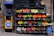 A colorful display of various fruits and vegetables arranged in black crates outside a store, next to a yellow vending machine with signage in a foreign script. The produce includes apples, oranges, lemons, peppers, potatoes, and onions with some water bottles nearby.