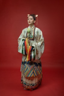 A person wearing intricate traditional attire stands against a deep red backdrop. The outfit is adorned with various colorful patterns and embroidery, including floral and geometric designs. The individual holds a white lotus flower and has ornate hair accessories with golden details.