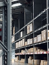 Shelving units filled with stacked boxes and pallets in a warehouse-like environment. The shelves are tall and extend into the distance, creating an orderly and spacious atmosphere. Overhead, industrial lighting and structural elements are visible.