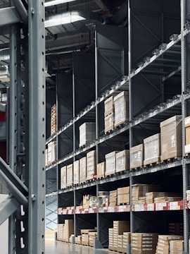 Shelving units filled with stacked boxes and pallets in a warehouse-like environment. The shelves are tall and extend into the distance, creating an orderly and spacious atmosphere. Overhead, industrial lighting and structural elements are visible.