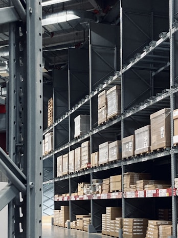 Shelving units filled with stacked boxes and pallets in a warehouse-like environment. The shelves are tall and extend into the distance, creating an orderly and spacious atmosphere. Overhead, industrial lighting and structural elements are visible.