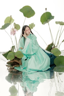 A person wearing a flowing, light turquoise robe sits among large lotus leaves and flowers. The setting appears serene and ethereal, with a reflective surface enhancing the tranquil atmosphere. The posture suggests a meditative or contemplative mood.