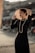 A person dressed elegantly in a black outfit with a pearl necklace, holding a wide-brimmed, straw hat against a blurred urban background. The scene is lit warmly, emphasizing the polished appearance and fashionable style.