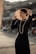 A person dressed elegantly in a black outfit with a pearl necklace, holding a wide-brimmed, straw hat against a blurred urban background. The scene is lit warmly, emphasizing the polished appearance and fashionable style.