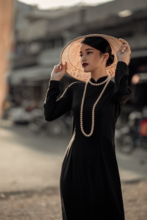 A person dressed elegantly in a black outfit with a pearl necklace, holding a wide-brimmed, straw hat against a blurred urban background. The scene is lit warmly, emphasizing the polished appearance and fashionable style.