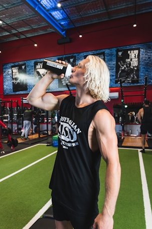 A person with light blonde hair is drinking from a shaker bottle in a gym. The gym has bright red walls and motivational posters on them. The floor features green turf, and there are various pieces of gym equipment in the background.