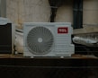 An outdoor air conditioning unit is mounted against a wall with a cracked and peeling surface. The unit displays a prominent TCL logo. Electrical cables are visible around the installation, with some pipes and barbed wire in the foreground, suggesting a secure or restricted area.