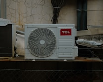 An outdoor air conditioning unit is mounted against a wall with a cracked and peeling surface. The unit displays a prominent TCL logo. Electrical cables are visible around the installation, with some pipes and barbed wire in the foreground, suggesting a secure or restricted area.