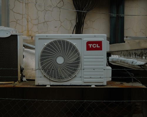 An outdoor air conditioning unit is mounted against a wall with a cracked and peeling surface. The unit displays a prominent TCL logo. Electrical cables are visible around the installation, with some pipes and barbed wire in the foreground, suggesting a secure or restricted area.
