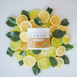 A container of 'Immunity' supplement is surrounded by slices of lemons and limes, along with fresh, green citrus leaves. The arrangement is carefully crafted for an appealing display on a flat white surface. The product label features images of citrus fruits and emphasizes health benefits.