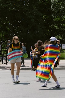 People are gathered outdoors, some wearing pride-themed clothing. A person holds a colorful sign with blue text, and another person is draped in a rainbow flag. The scene appears lively with individuals engaging in conversation. Bicycles and trees are visible in the background, adding to the festive and inclusive atmosphere.