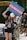 A person holds up a colorful sign advocating for accessible transgender healthcare, standing in front of a vehicle with others nearby. They are wearing a cap, floral shirt, and shorts, and appear to be participating in a public demonstration, with a park gate visible in the background.