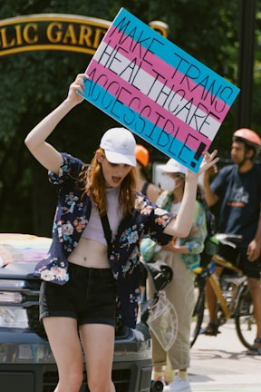 A person holds up a colorful sign advocating for accessible transgender healthcare, standing in front of a vehicle with others nearby. They are wearing a cap, floral shirt, and shorts, and appear to be participating in a public demonstration, with a park gate visible in the background.