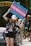 A person holds up a colorful sign advocating for accessible transgender healthcare, standing in front of a vehicle with others nearby. They are wearing a cap, floral shirt, and shorts, and appear to be participating in a public demonstration, with a park gate visible in the background.