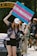 A person holds up a colorful sign advocating for accessible transgender healthcare, standing in front of a vehicle with others nearby. They are wearing a cap, floral shirt, and shorts, and appear to be participating in a public demonstration, with a park gate visible in the background.