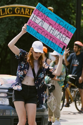 A person holds up a colorful sign advocating for accessible transgender healthcare, standing in front of a vehicle with others nearby. They are wearing a cap, floral shirt, and shorts, and appear to be participating in a public demonstration, with a park gate visible in the background.