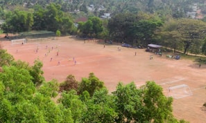 An aerial view of a football field surrounded by lush green trees. Players in colorful uniforms are actively engaged in a game, with spectators watching from the sidelines under shaded areas. The field is bordered by a netted fence on one side, and the area has a natural, rustic feel due to the visible soil.
