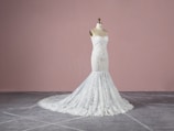 A strapless, white lace wedding gown displayed on a dress form. The gown features intricate detailing with a form-fitting bodice that extends into a full-length skirt with a train. The background is a plain, muted pink wall and the floor is grey with a subtle pattern.