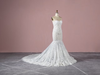 A strapless, white lace wedding gown displayed on a dress form. The gown features intricate detailing with a form-fitting bodice that extends into a full-length skirt with a train. The background is a plain, muted pink wall and the floor is grey with a subtle pattern.