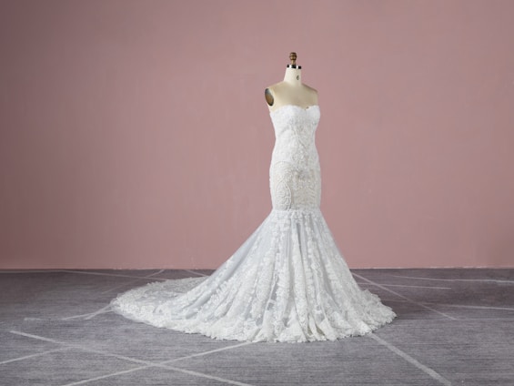A strapless, white lace wedding gown displayed on a dress form. The gown features intricate detailing with a form-fitting bodice that extends into a full-length skirt with a train. The background is a plain, muted pink wall and the floor is grey with a subtle pattern.