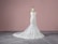A strapless, white lace wedding gown displayed on a dress form. The gown features intricate detailing with a form-fitting bodice that extends into a full-length skirt with a train. The background is a plain, muted pink wall and the floor is grey with a subtle pattern.
