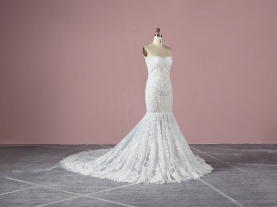A strapless, white lace wedding gown displayed on a dress form. The gown features intricate detailing with a form-fitting bodice that extends into a full-length skirt with a train. The background is a plain, muted pink wall and the floor is grey with a subtle pattern.