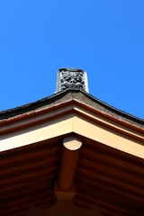 A traditional rooftop featuring intricate architectural details, with a decorative emblem positioned at the peak against a clear blue sky.