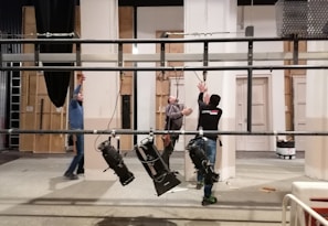 Three individuals are working on a set, adjusting lighting equipment suspended from a metal rig. The background includes panels that resemble part of a stage set, with a ladder visible on the left. The environment suggests an indoor studio setting.