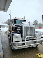 A large Kenworth semi-truck is parked at a service station on a wet concrete surface. The truck has a chrome grille and is equipped with visible pipes on its sides. There is a water hose lying on the ground nearby, and the background includes a building and some leafless trees on a cloudy day.