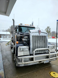 A large Kenworth semi-truck is parked at a service station on a wet concrete surface. The truck has a chrome grille and is equipped with visible pipes on its sides. There is a water hose lying on the ground nearby, and the background includes a building and some leafless trees on a cloudy day.