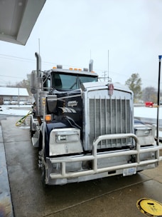 A large Kenworth semi-truck is parked at a service station on a wet concrete surface. The truck has a chrome grille and is equipped with visible pipes on its sides. There is a water hose lying on the ground nearby, and the background includes a building and some leafless trees on a cloudy day.