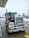 A large Kenworth semi-truck is parked at a service station on a wet concrete surface. The truck has a chrome grille and is equipped with visible pipes on its sides. There is a water hose lying on the ground nearby, and the background includes a building and some leafless trees on a cloudy day.