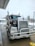 A large Kenworth semi-truck is parked at a service station on a wet concrete surface. The truck has a chrome grille and is equipped with visible pipes on its sides. There is a water hose lying on the ground nearby, and the background includes a building and some leafless trees on a cloudy day.