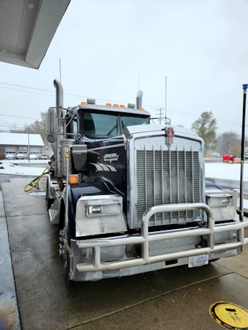 A large Kenworth semi-truck is parked at a service station on a wet concrete surface. The truck has a chrome grille and is equipped with visible pipes on its sides. There is a water hose lying on the ground nearby, and the background includes a building and some leafless trees on a cloudy day.