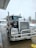 A large Kenworth semi-truck is parked at a service station on a wet concrete surface. The truck has a chrome grille and is equipped with visible pipes on its sides. There is a water hose lying on the ground nearby, and the background includes a building and some leafless trees on a cloudy day.