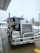 A large Kenworth semi-truck is parked at a service station on a wet concrete surface. The truck has a chrome grille and is equipped with visible pipes on its sides. There is a water hose lying on the ground nearby, and the background includes a building and some leafless trees on a cloudy day.
