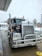 A large Kenworth semi-truck is parked at a service station on a wet concrete surface. The truck has a chrome grille and is equipped with visible pipes on its sides. There is a water hose lying on the ground nearby, and the background includes a building and some leafless trees on a cloudy day.