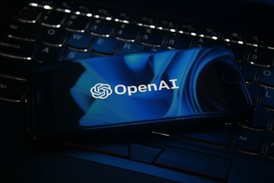 A smartphone displaying the OpenAI logo rests on a laptop keyboard. The screen features a blue abstract design, and the keyboard is visible beneath with dimly lit keys.