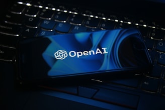 A smartphone displaying the OpenAI logo rests on a laptop keyboard. The screen features a blue abstract design, and the keyboard is visible beneath with dimly lit keys.