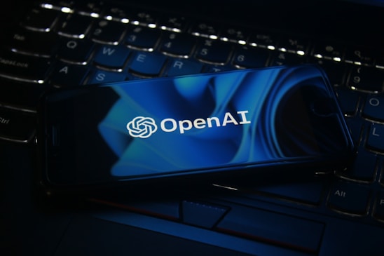 A smartphone displaying the OpenAI logo rests on a laptop keyboard. The screen features a blue abstract design, and the keyboard is visible beneath with dimly lit keys.