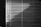 Horizontal blinds cover a window, with light creating a pattern of alternating light and dark stripes. The pull cords hang on the left side, casting small shadows.