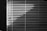 Horizontal blinds cover a window, with light creating a pattern of alternating light and dark stripes. The pull cords hang on the left side, casting small shadows.