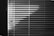Horizontal blinds cover a window, with light creating a pattern of alternating light and dark stripes. The pull cords hang on the left side, casting small shadows.