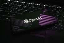 A smartphone displaying the OpenAI logo is resting on a laptop keyboard. The phone screen reflects purple and white light patterns, adding a modern and tech-focused ambiance.