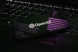 A smartphone displaying the OpenAI logo is resting on a laptop keyboard. The phone screen reflects purple and white light patterns, adding a modern and tech-focused ambiance.