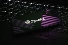 A smartphone displaying the OpenAI logo is resting on a laptop keyboard. The phone screen reflects purple and white light patterns, adding a modern and tech-focused ambiance.